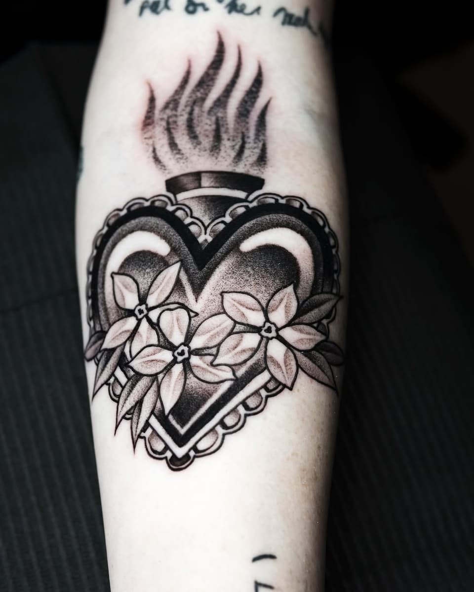 Sacred heart tattoo with ornamental frame, flames, and white flowers on the upper arm in black and grey ink.