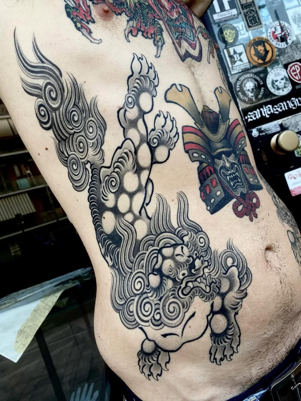 Large ornate lion with spiraling mane and decorative paws on torso, accompanied by smaller colorful demon mask tattoo on upper chest.
