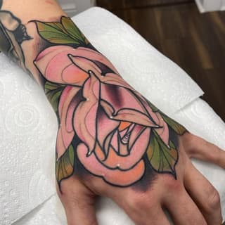 Colorful multi-layered pink and orange flower with green leaves tattooed on the back of a hand, featuring bold black outlines.