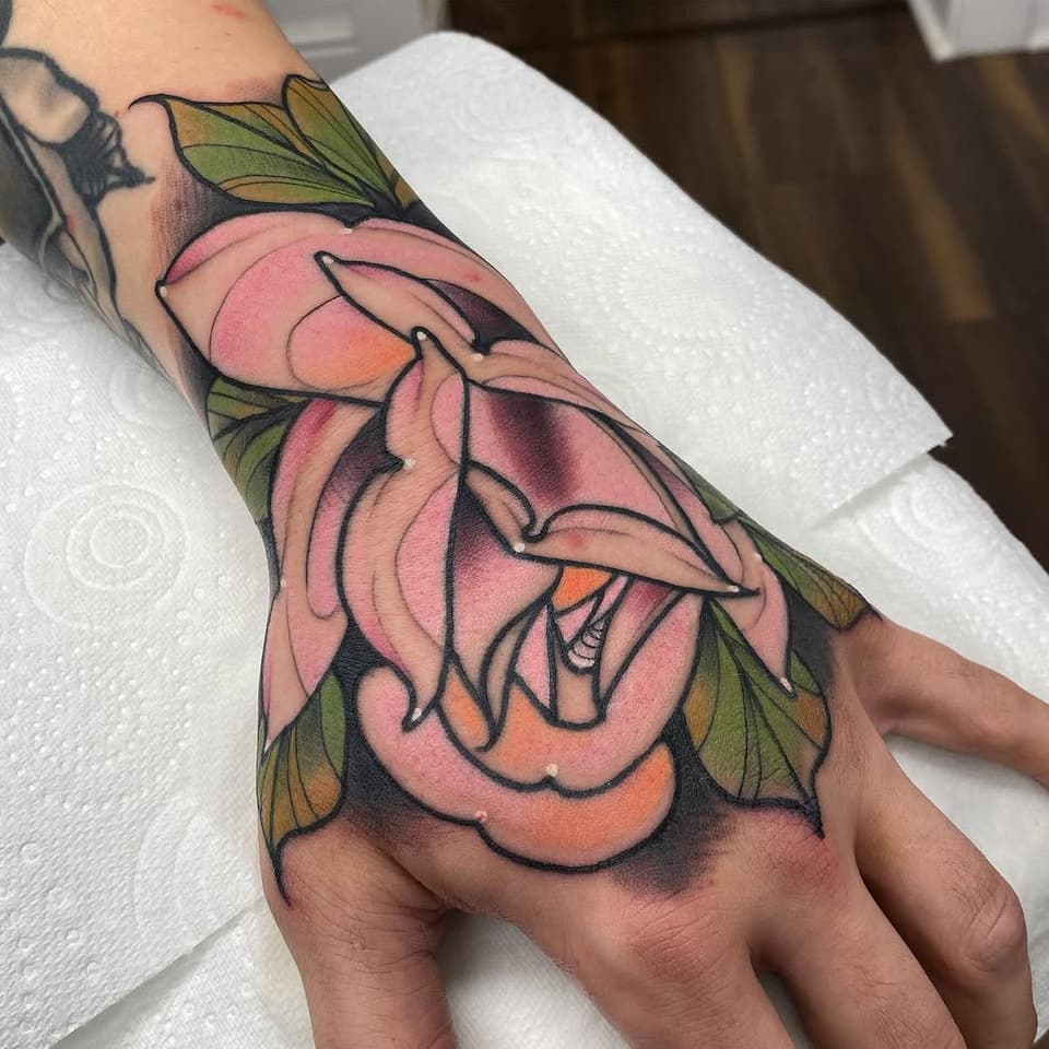 Colorful multi-layered pink and orange flower with green leaves tattooed on the back of a hand, featuring bold black outlines.