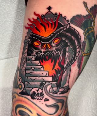 Colorful skull tattoo with red and orange flames erupting from the crown, stone structure body, and decorative elements on the arm.