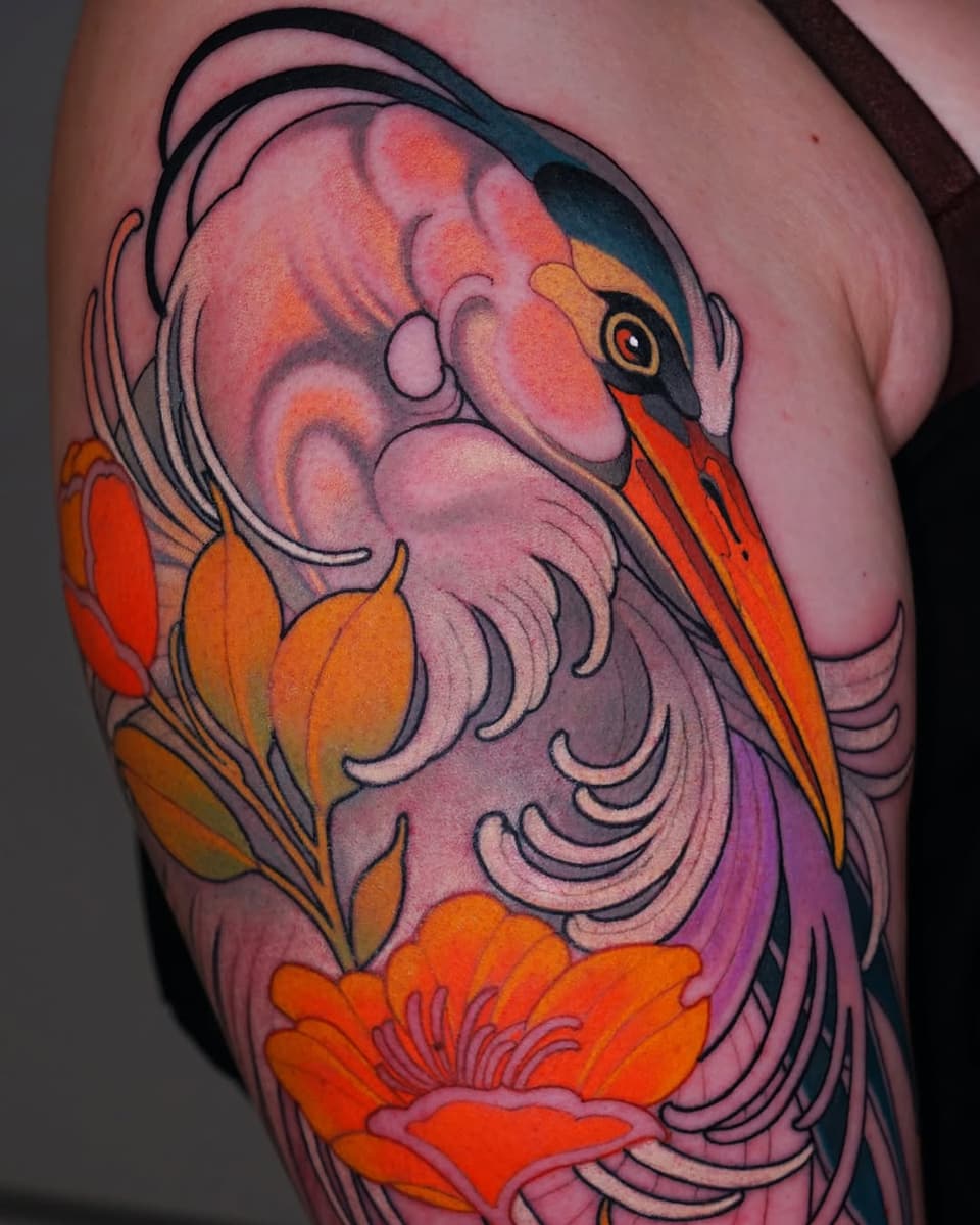 Colorful bird tattoo on upper arm featuring pink, orange, and red plumage with detailed feather work and orange flowers below.