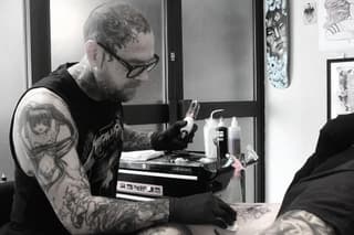 Tattoo artist wearing glasses and black gloves works on a client's arm with a tattoo machine in a studio setting.