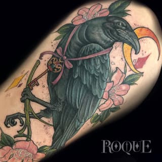 Dark raven holding an ornate key with pink flowers and a crescent moon on the upper arm, rendered in black, pink, green, and gold.
