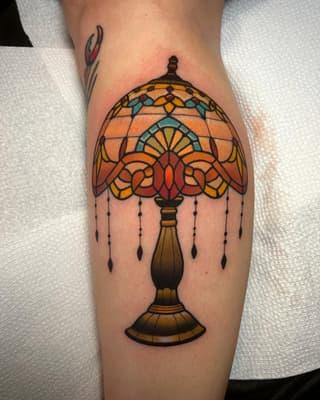 Stained glass lamp tattoo with orange, yellow, and teal colors on the calf, featuring a decorative base and hanging beads.