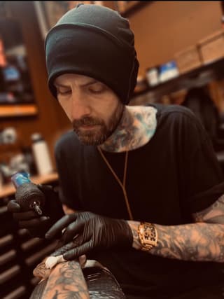 Tattoo artist wearing black beanie and gloves works on client's arm displaying blue and black ink designs with gold watch visible.