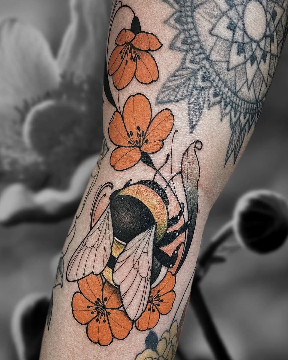 Colorful tattoo on an arm featuring orange flowers, a detailed bee with black and gold wings, green leaves, and surrounding botanical elements.