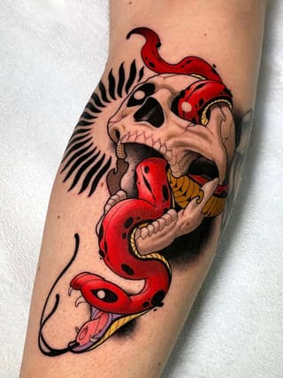 Skull with wings and red serpent wrapping around it, featuring gold accents, tattooed on the upper arm.