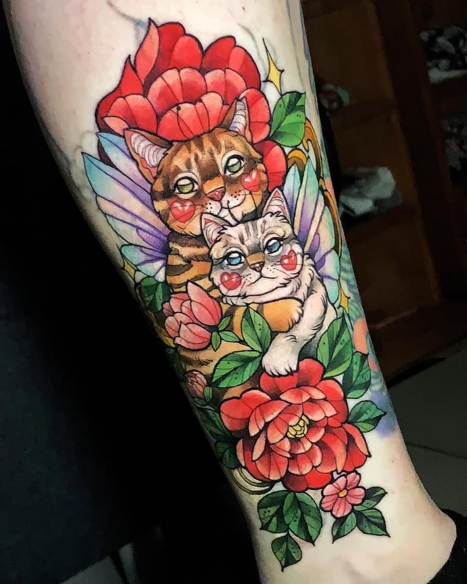 Colorful tattoo on a calf featuring two winged cats with heart-marked faces surrounded by red and pink flowers with green leaves.