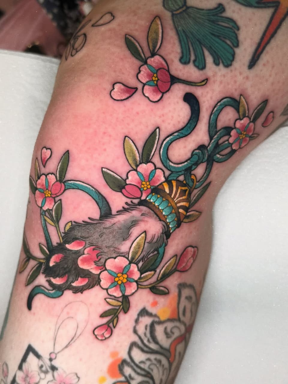 A colorful arm tattoo featuring a deer surrounded by pink flowers, teal vines, and a butterfly, with green leaves and yellow accents throughout.
