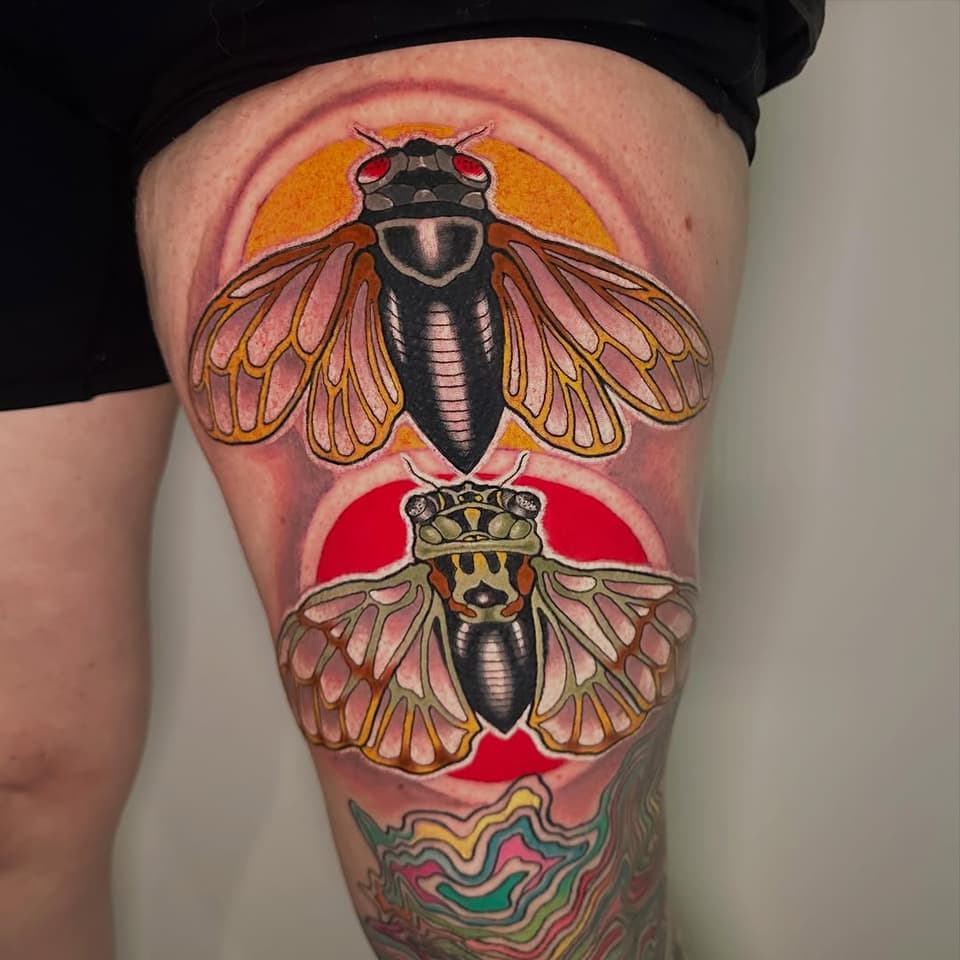 Colorful thigh tattoo featuring two insects with wings, crowns, and circular backgrounds in orange and red, with decorative wavy patterns below.