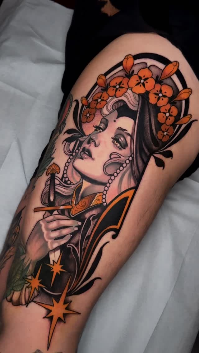 Woman's upper arm displaying a portrait tattoo of a face with orange flowers in hair, pearl necklace, and decorative elements in orange and black ink.