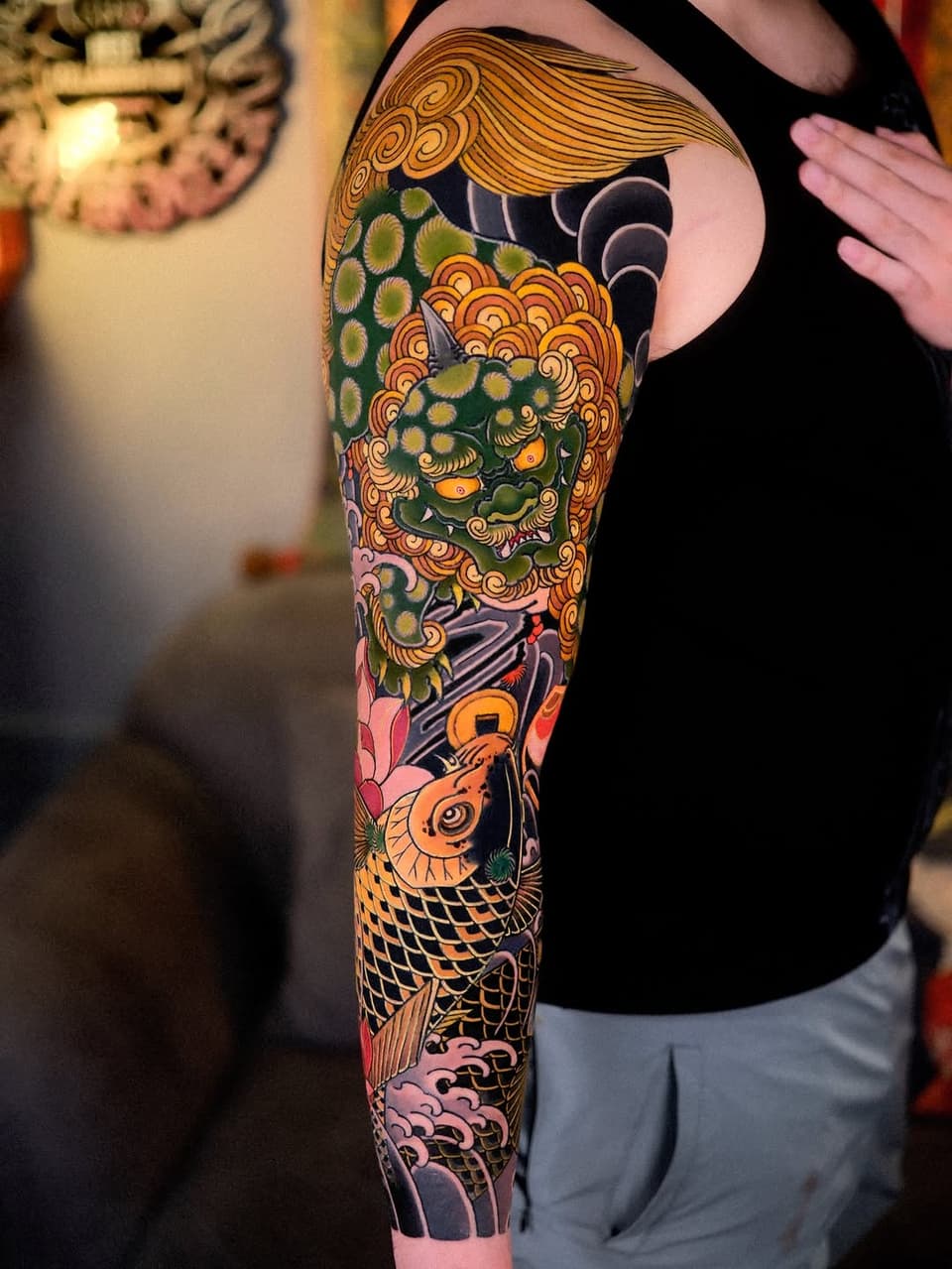 Full-sleeve tattoo featuring a green demon mask with ornate golden scrollwork, orange koi fish, pink flowers, and decorative patterns in vibrant colors on the arm.