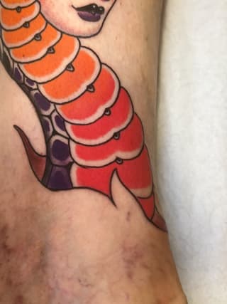 Mermaid tail tattoo in orange, red, and purple gradients with black outlines on the lower arm, featuring scaled details and a female face above.
