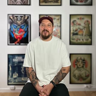 Tattooed man with facial hair wearing white t-shirt and burgundy cap, seated before framed artworks displaying colorful figure illustrations on white wall.