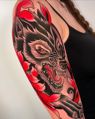 Large demon face tattoo on upper arm in red and black ink with detailed features, sharp teeth, red eyes, and red floral elements surrounding the design.