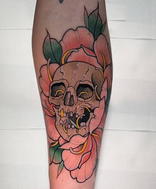 Skull surrounded by pink flowers with green leaves and gold accents tattooed on the calf.