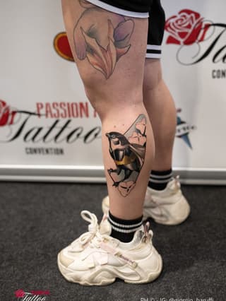 Leg tattoo featuring a bat with spread wings in black and tan tones above a colorful flower design on the thigh, displayed in a studio setting.