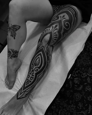 Large ornamental leg sleeve tattoo featuring symmetrical geometric and floral patterns in black ink on pale skin, displayed in black and white photography.
