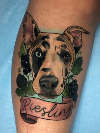 Portrait tattoo of a dog with black and tan markings, grapes and vines in green, and a pink banner reading "Blessing" on the calf.