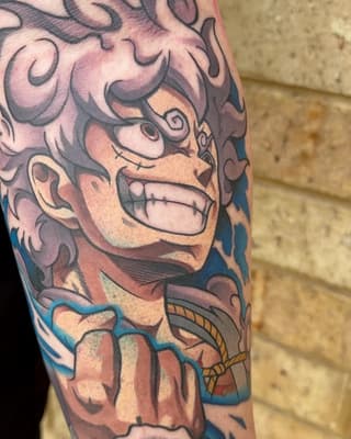 Colorful demon face tattoo on upper arm with ornamental scrollwork, featuring red, blue, and gold accents on pale skin against a brick wall.