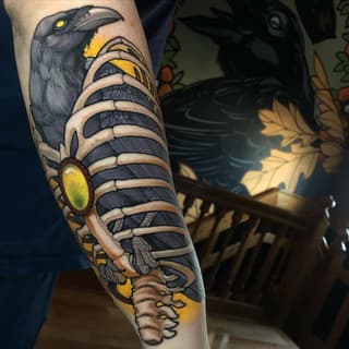 Colorful arm tattoo featuring a black raven with yellow eye at top, striped caterpillar body with yellow spots, and decorative roots at bottom in yellow, black, and tan.