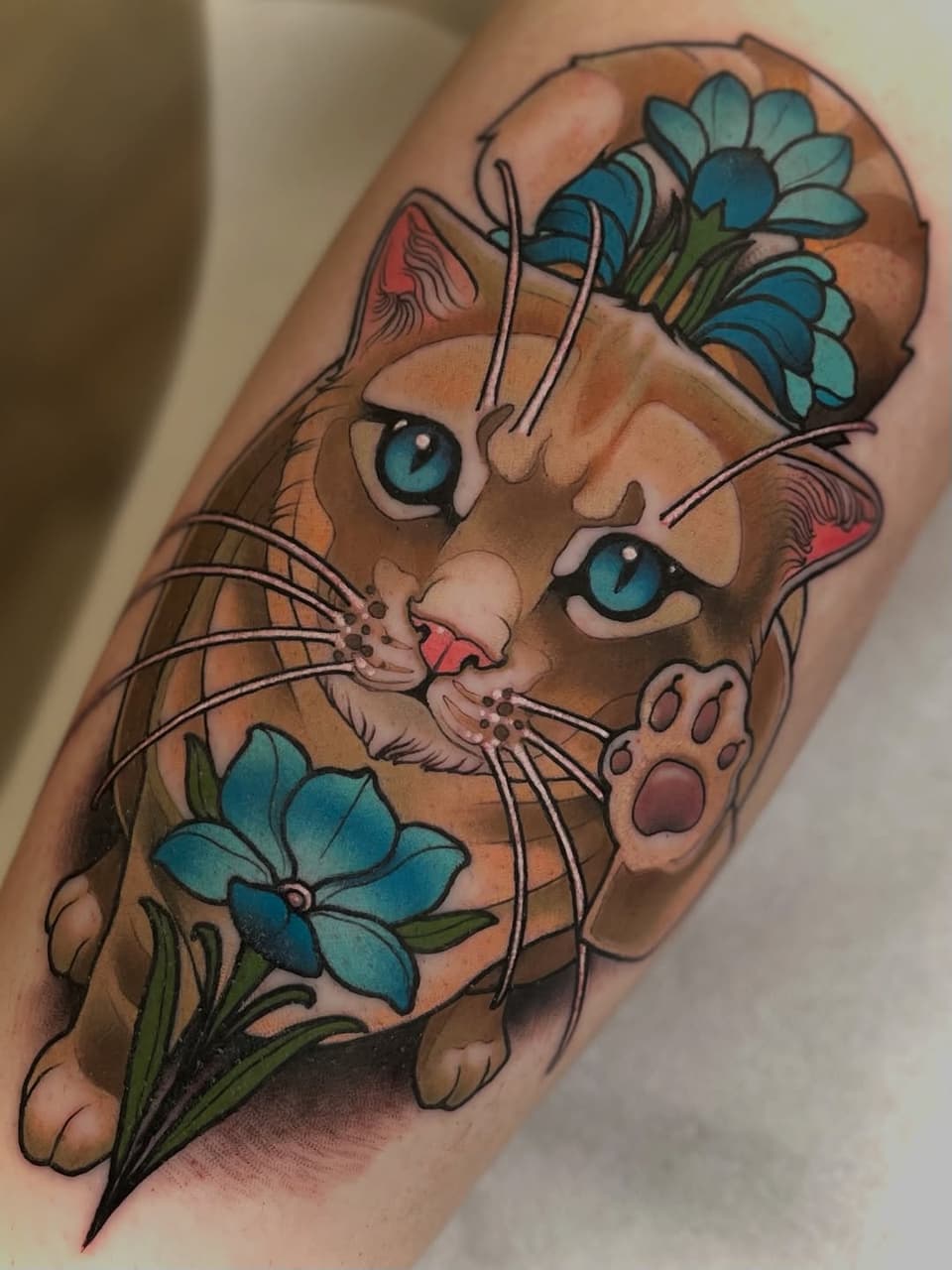 Orange tabby cat with blue eyes and pink nose surrounded by blue flowers with green stems, tattooed on upper arm.