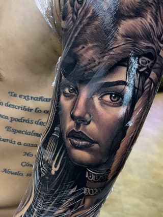 Portrait tattoo of a woman's face with long hair, decorative neck ornaments, and Spanish text on the upper arm, done in black ink on skin.