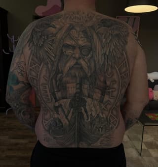 Large back tattoo featuring a bearded man with an eye patch and winged figures, surrounded by ornate geometric patterns and runic symbols in black and gray ink.