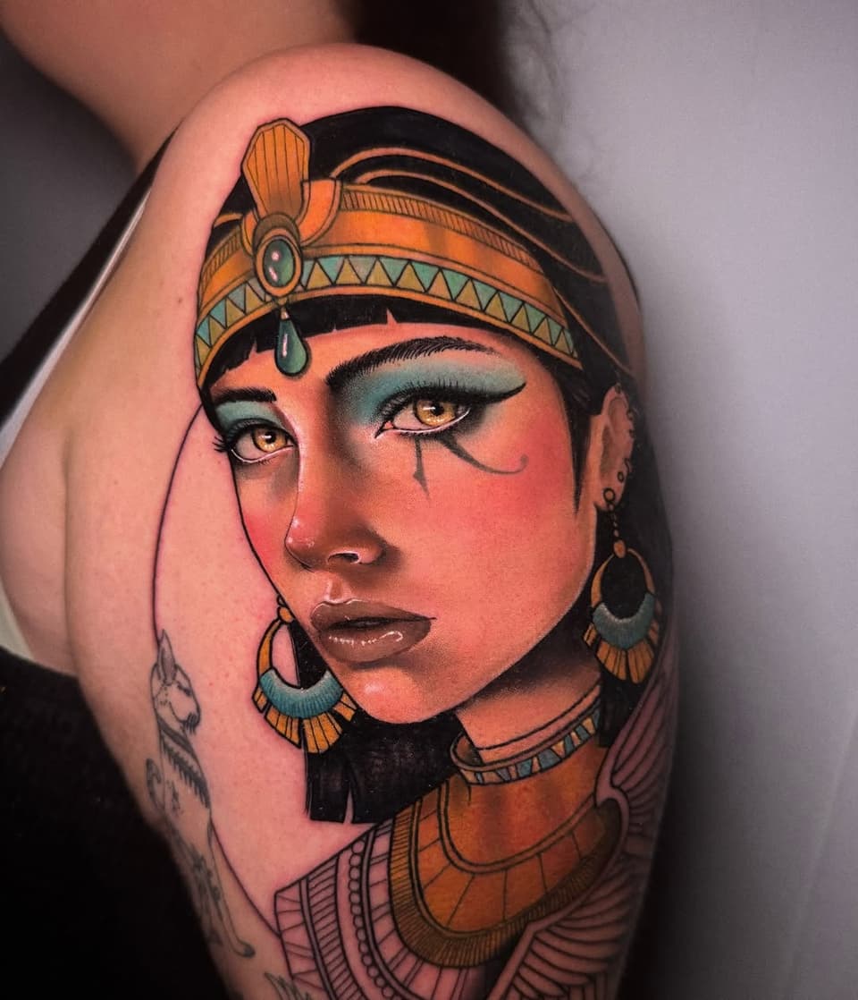 Colorful shoulder tattoo depicting an Egyptian-inspired female figure with ornate headdress, jewelry, and Egyptian Eye of Horus makeup in orange, teal, and gold tones.