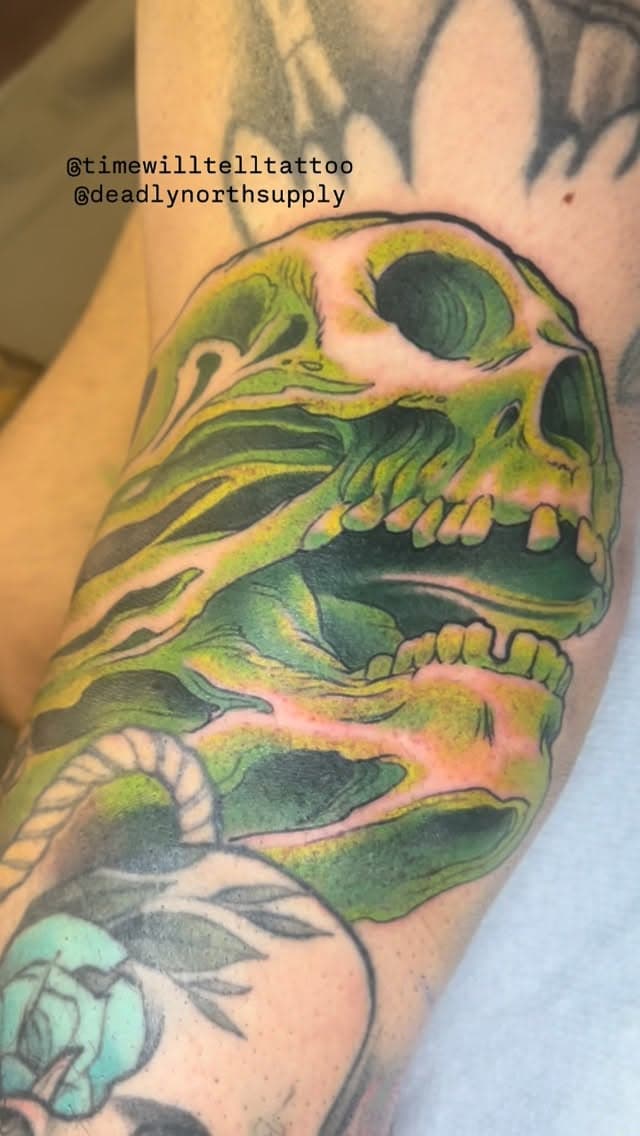 Stacked green and yellow skull design with detailed bone structure and shading on upper arm, with partial floral elements visible below.
