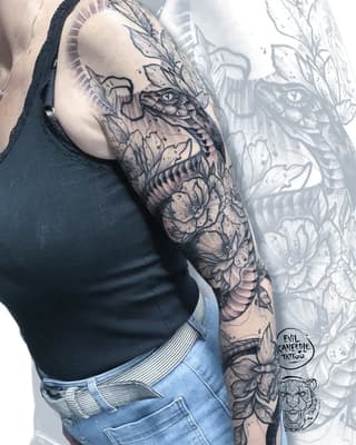 Full arm sleeve tattoo featuring a dragon head, flowers, and botanical elements in black and gray ink on a person's shoulder and upper arm.