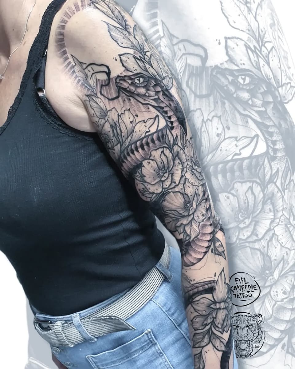 Full arm sleeve tattoo featuring a dragon head, flowers, and botanical elements in black and gray ink on a person's shoulder and upper arm.