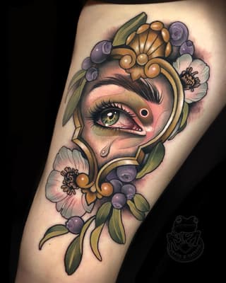 Colorful eye tattoo with ornamental gold frames, purple berries, white flowers, and green leaves on the forearm.