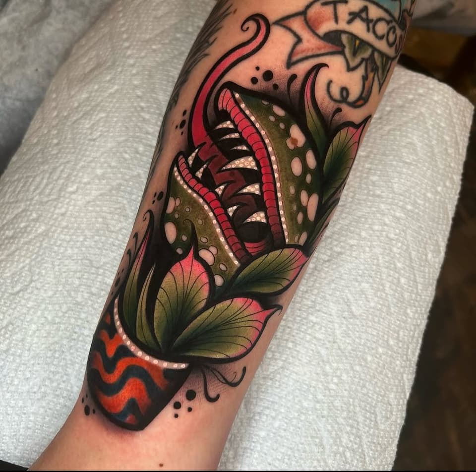 Colorful carnivorous plant tattoo on forearm featuring red petals, green leaves, and open mouth with teeth, surrounded by decorative elements.