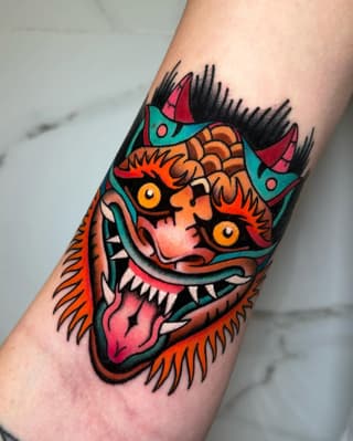 Colorful demon mask tattoo on forearm featuring orange, red, turquoise, and yellow with prominent eyes, horns, and sharp teeth.