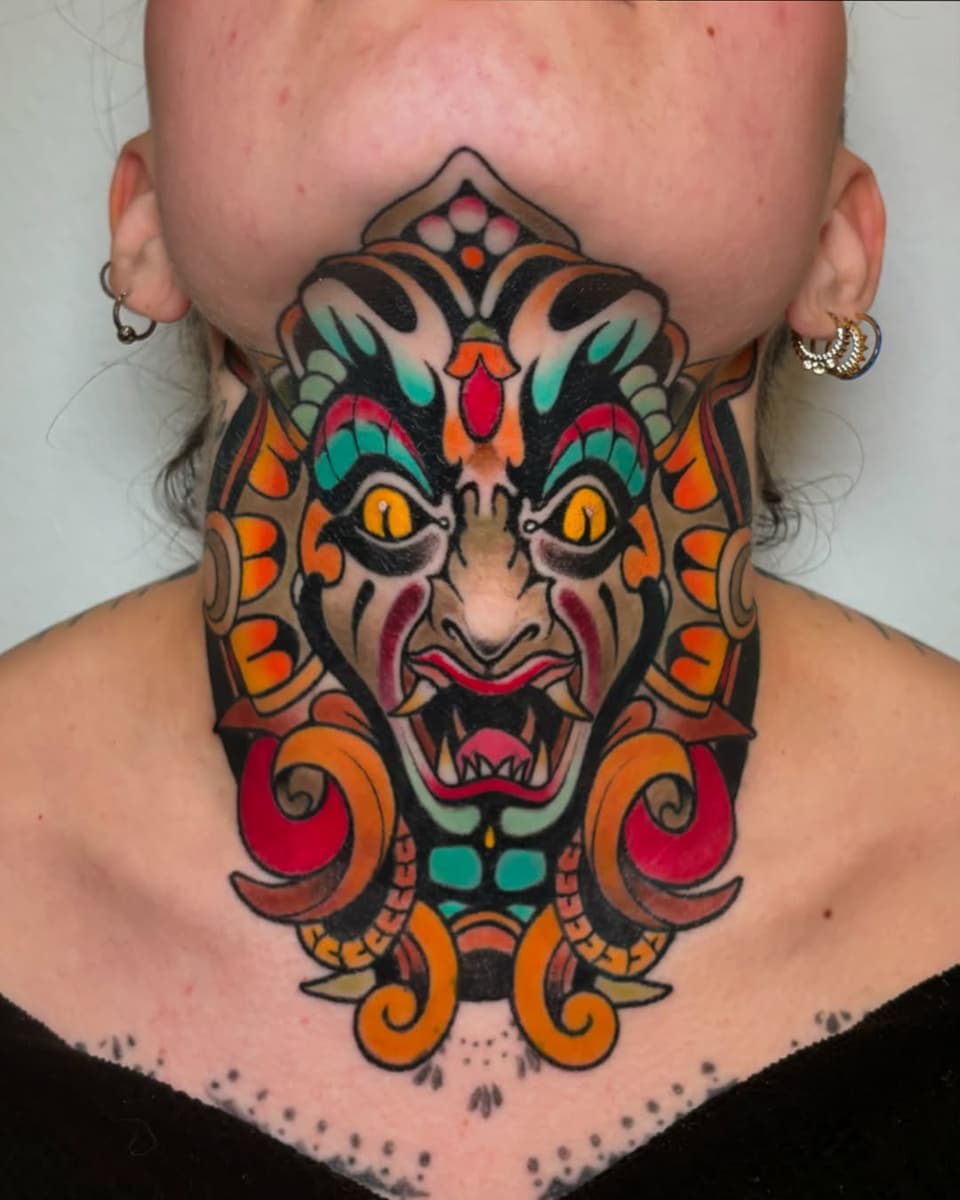 Colorful demon mask tattoo on neck and chest featuring golden eyes, red mouth, orange scrollwork, and turquoise accents with symmetrical design.
