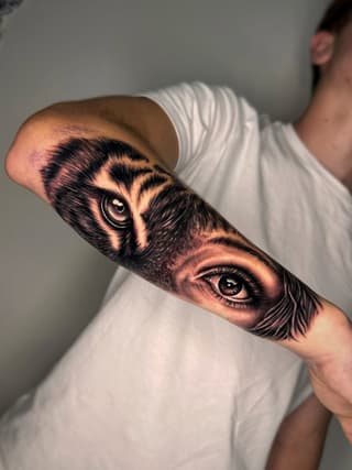 Two owl and human eyes tattooed on a forearm in black and brown ink, displayed on a person wearing a white shirt.