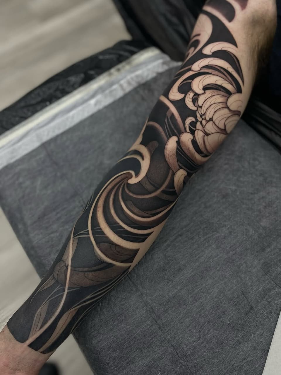 Ornate scrolling sleeve tattoo in black ink with tan and brown shading, featuring flowing baroque-inspired flourishes wrapped around the forearm and upper arm.