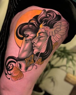Colorful shoulder tattoo featuring a profile of a woman with elaborate headdress, wings, pearl necklace, and ornamental details in orange, pink, and black.