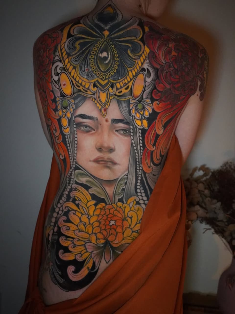 Large back tattoo featuring a portrait of a figure wearing ornate headwear and jewelry, surrounded by decorative floral motifs in orange, gold, red, and black.