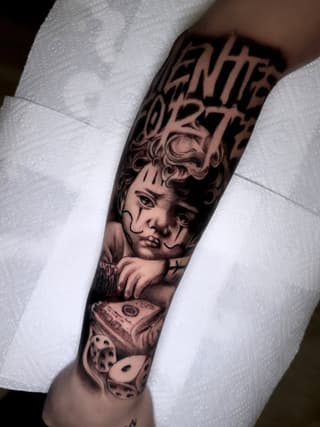 Detailed cherub face tattoo on forearm with text above, money and dice below, rendered in black and grey ink on pale skin.