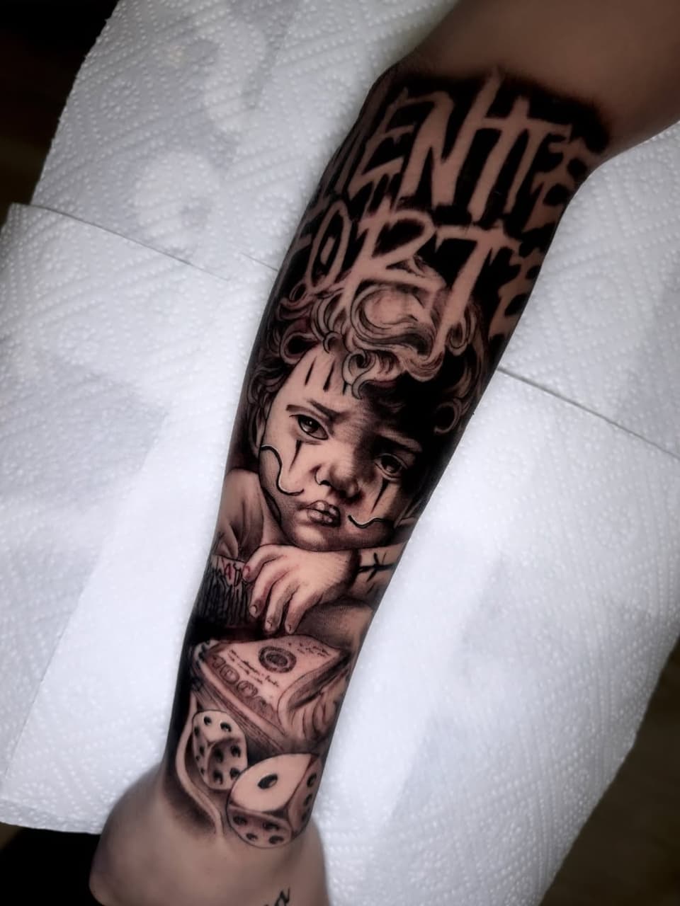 Detailed cherub face tattoo on forearm with text above, money and dice below, rendered in black and grey ink on pale skin.