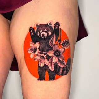 Red panda with raised paws holding pink flowers against a bright orange circular background, tattooed on the thigh.