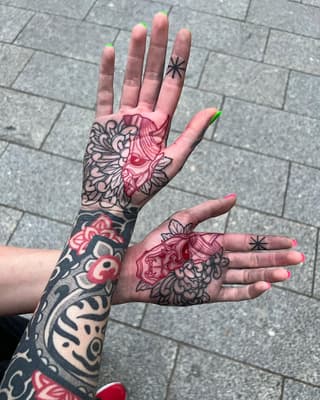 Two hands displaying palm tattoos featuring ornate floral and mask designs in black and red ink, with matching star tattoos on the fingers.