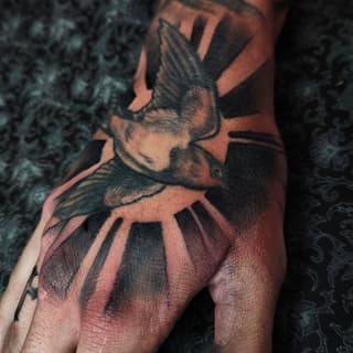 A raven with spread wings and detailed feather work in black and gray ink on the back of a hand, featuring shading and depth.