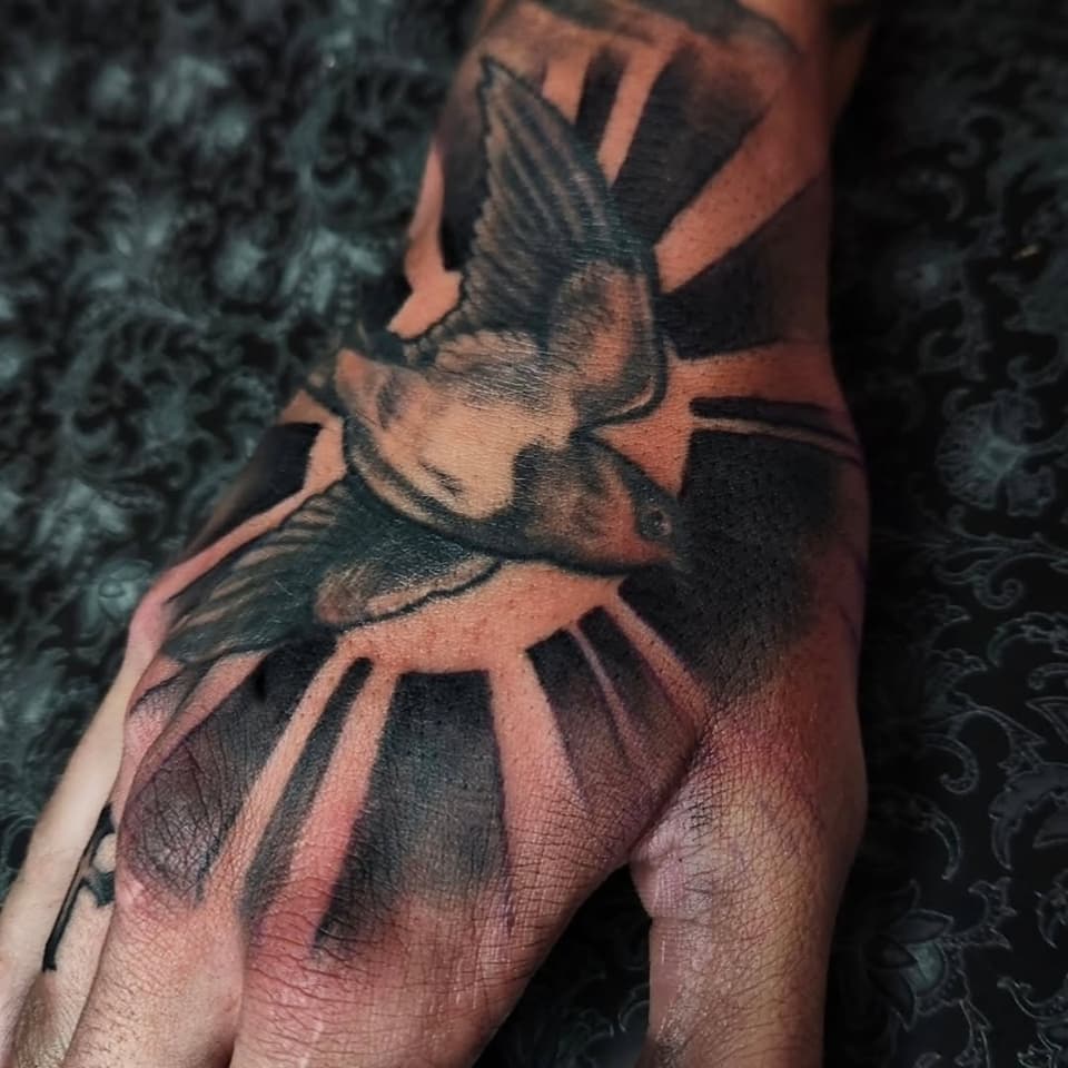 A raven with spread wings and detailed feather work in black and gray ink on the back of a hand, featuring shading and depth.