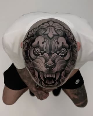 Overhead view of a detailed demon or oni mask tattoo on a person's scalp, featuring intricate shading in grays and blacks with prominent eyes and bared teeth.