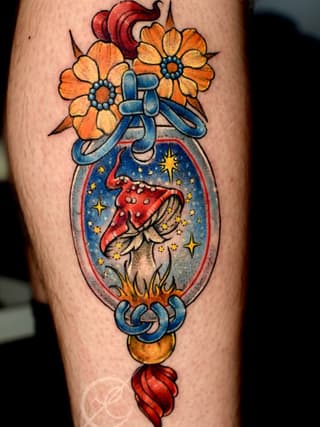 Colorful tattoo on a leg featuring orange flowers with a blue bow above an ornate oval frame containing a red mushroom surrounded by stars and celestial elements.