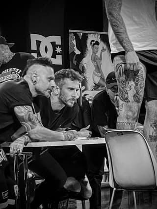 Tattoo artist displays arm sleeve featuring a large dragon design in black ink during a convention panel discussion.